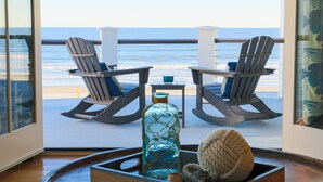 Un balcon avec deux fauteuils Adirondack donnant sur l’océan, une petite table avec une bouteille et une pelote de laine, et une vue sur la plage.
