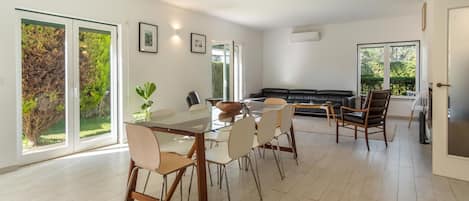 A spacious living room with a glass dining table, comfortable seating, and large windows offering a view of greenery.