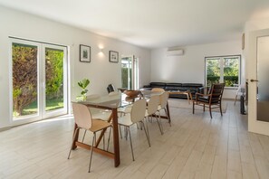 A spacious living room with a glass dining table, comfortable seating, and large windows offering a view of greenery. - LVPremier LA1- heated pool, Jacuzzi, AC, garden (Sesimbra)