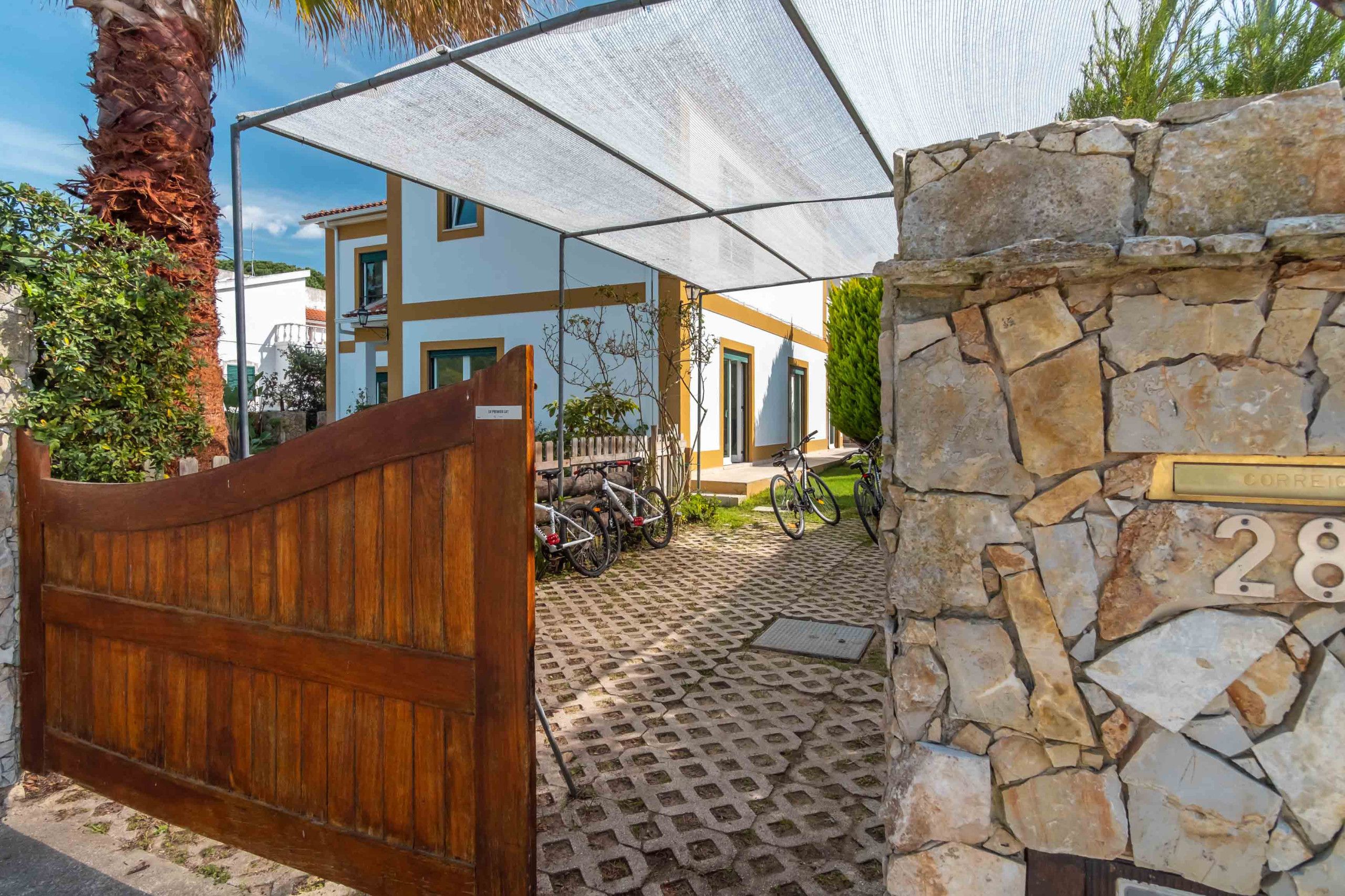 A gated entrance with a stone wall and a numbered plaque, a covered patio area with bicycles, and a white building with a yellow trim in the background.
