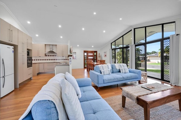 A modern kitchen with white cabinets and a central island, a living area with blue sofas and a wooden coffee table, and large windows offering a view of the outdoors.
