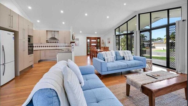 A modern kitchen with white cabinets and a central island, a living area with blue sofas and a wooden coffee table, and large windows offering a view of the outdoors. - Gathering of 8 Holiday House (Agnes Water)