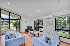 A spacious living room with two light blue sofas, a wooden coffee table, and a TV on a stand. Large windows offer views of a fenced outdoor area with greenery.