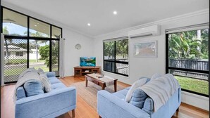 A spacious living room with two light blue sofas, a wooden coffee table, and a TV on a stand. Large windows offer views of a fenced outdoor area with greenery. - Gathering of 8 Holiday House (Agnes Water)