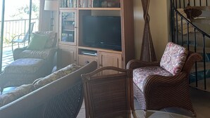 Un salon chaleureux avec un plafond haut, un meuble de rangement en bois, deux tableaux encadrés, des meubles en osier et un escalier.