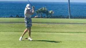 Une personne jouant au golf par une journée ensoleillée, avec une vue dégagée sur l’océan et des palmiers en arrière-plan.