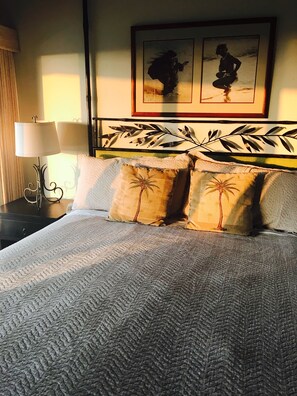 Un lit bien fait, recouvert d’une couverture à motifs, avec deux coussins en forme de palmier, une tête de lit décorative en métal et un tableau encadré au-dessus du lit.