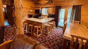 Ein Blockhaus-Interieur mit einer zentralen Barinsel, Holzstühlen, einem Esstisch und einem großen Fenster.