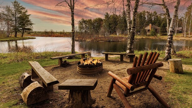 Ein Campingplatz am Seeufer mit Feuerstelle, Holzbänken und einem Stuhl.