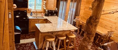 Interior de uma cabana de madeira com cozinha, bancada de granito, banquetas de madeira e mesa de jantar.