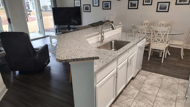 A modern kitchen with granite countertops, a built-in sink, and white cabinetry. There is a dining area with a glass table and white chairs.