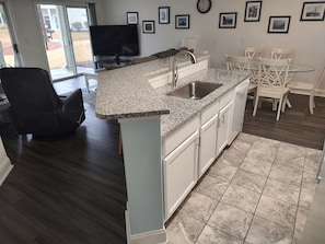Cozinha moderna com bancadas de granito, pia embutida e armários brancos. Há uma área de jantar com mesa de vidro e cadeiras brancas.