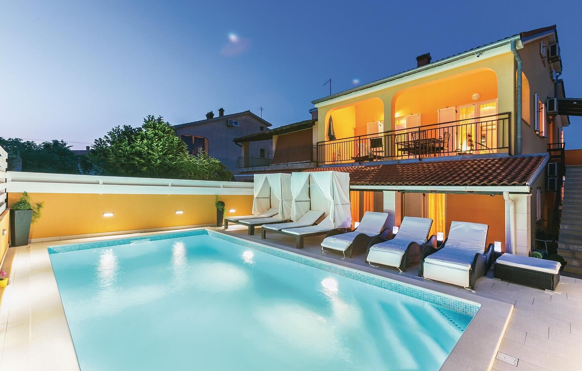 A swimming pool with lounge chairs and umbrellas, a balcony with outdoor seating, and a building with warm lighting.