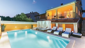 En swimmingpool med liggestole og parasoller, en balkon med udendørs møbler, og en bygning med varm belysning.