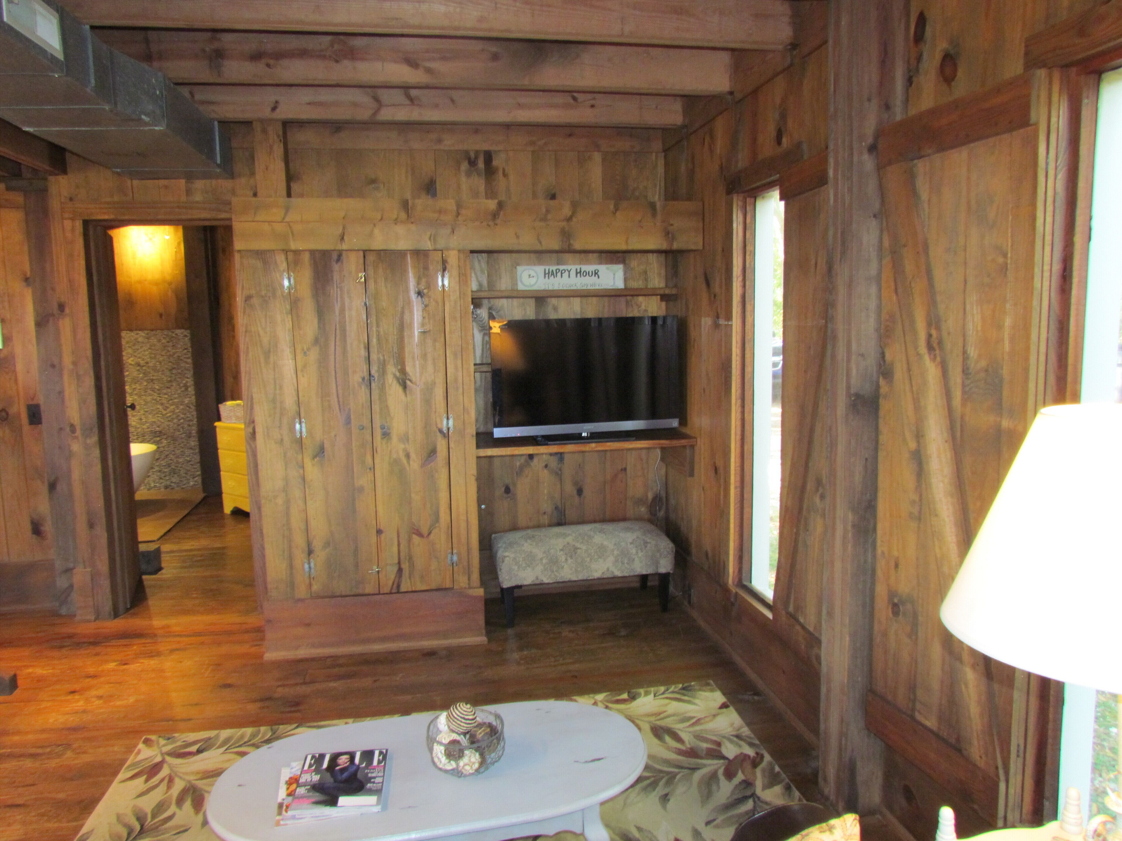 Interior de una cabaña de madera con un televisor montado en la pared, una pequeña zona de estar y una puerta que da a un baño.
