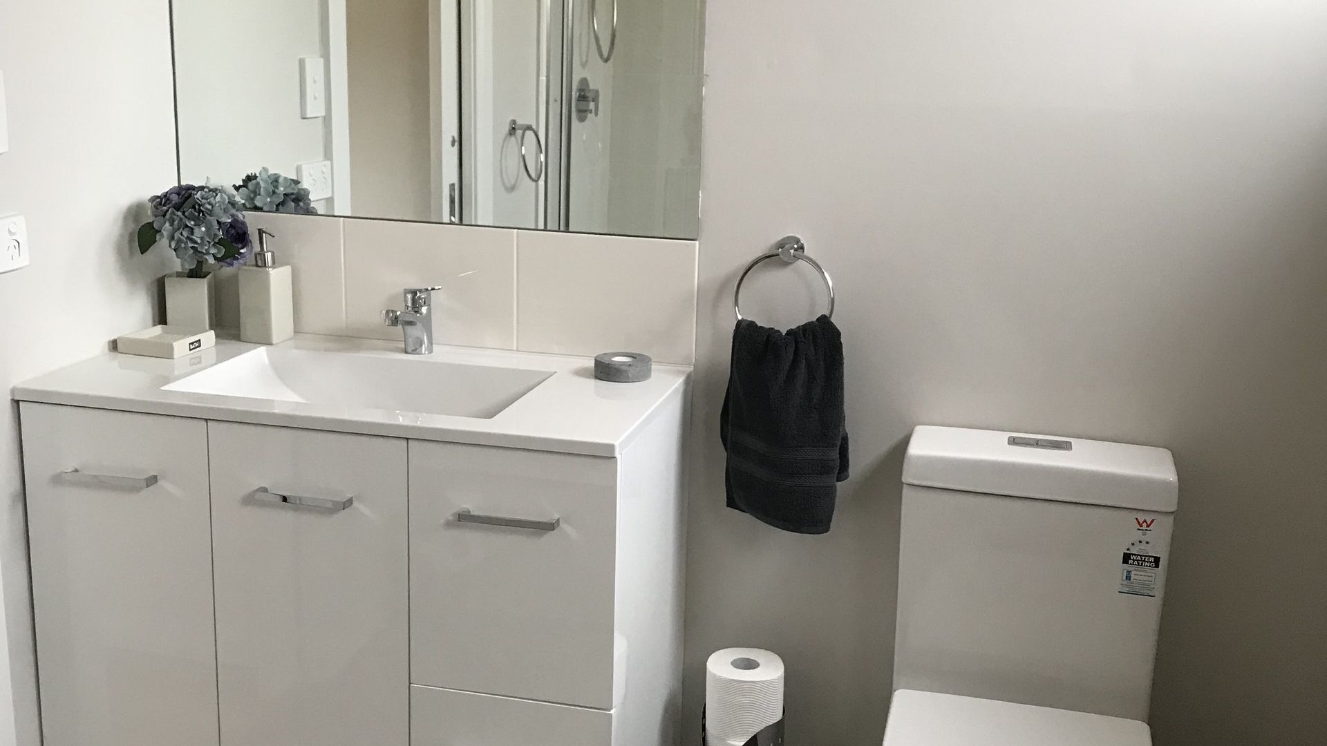 Ein Badezimmer mit Waschbecken, Spiegel und Toilette. Ein Handtuchhalter mit einem Handtuch und eine Toilettenpapierrolle auf dem Boden.