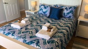 A neatly made bed with a tropical leaf pattern bedspread, two folded towels, and a blue pillow.