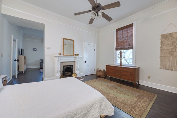 A bedroom with a large bed, a fireplace, a wooden dresser, a ceiling fan, and a window with blinds. - Dimon Cottage, Suite A (Columbus)