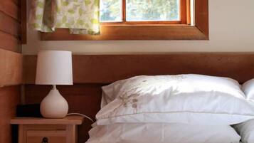 A bedroom with a wooden headboard, a bedside lamp, a nightstand with a drawer, and a window with a view of greenery.