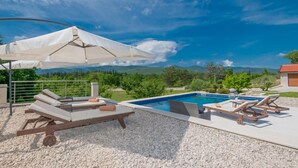 A pool area with lounge chairs and umbrellas, surrounded by a gravel deck and a view of green hills and mountains.