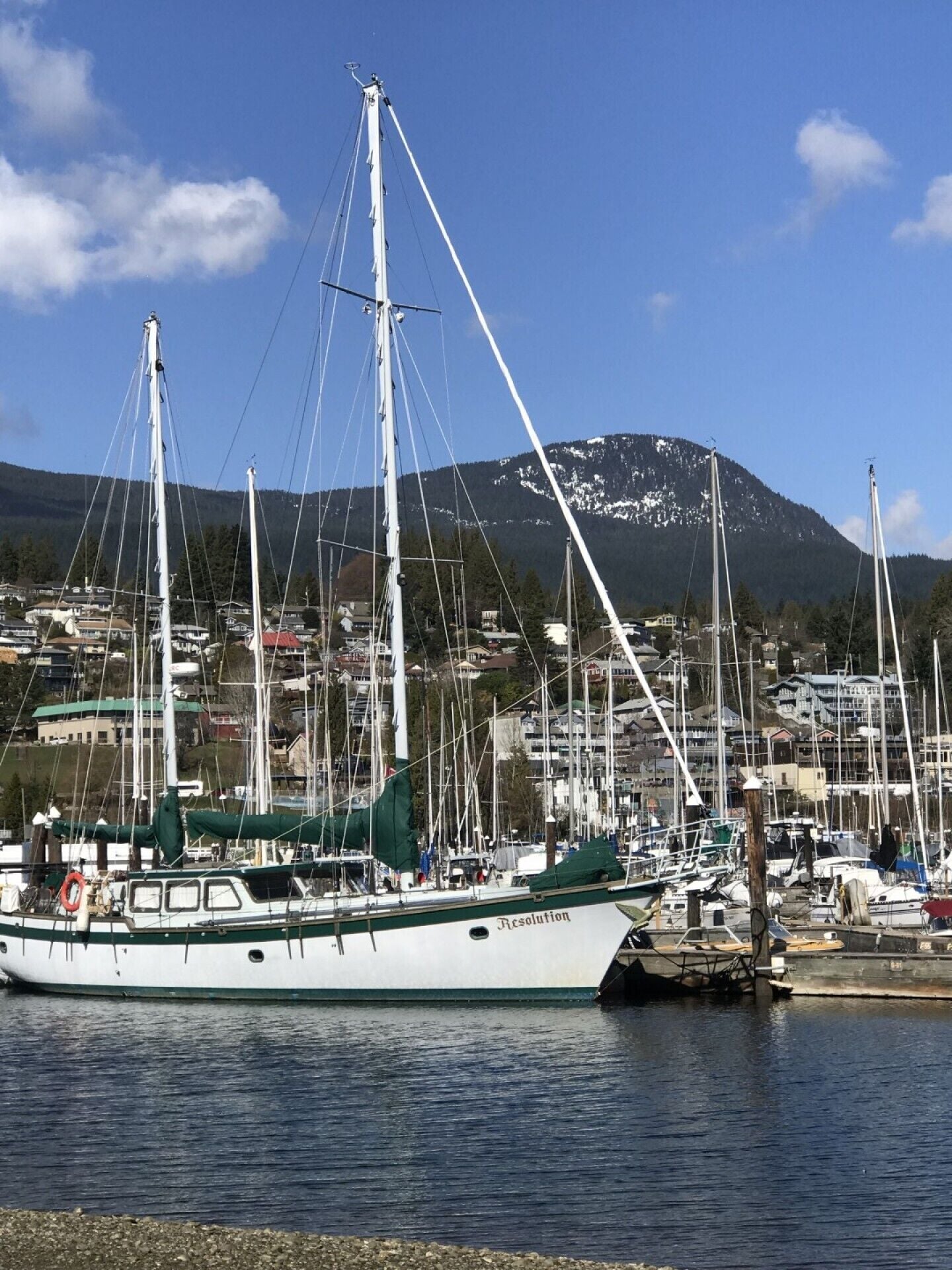 Un port de plaisance où sont amarrés plusieurs voiliers, avec une chaîne de montagnes en arrière-plan et une zone résidentielle au loin.