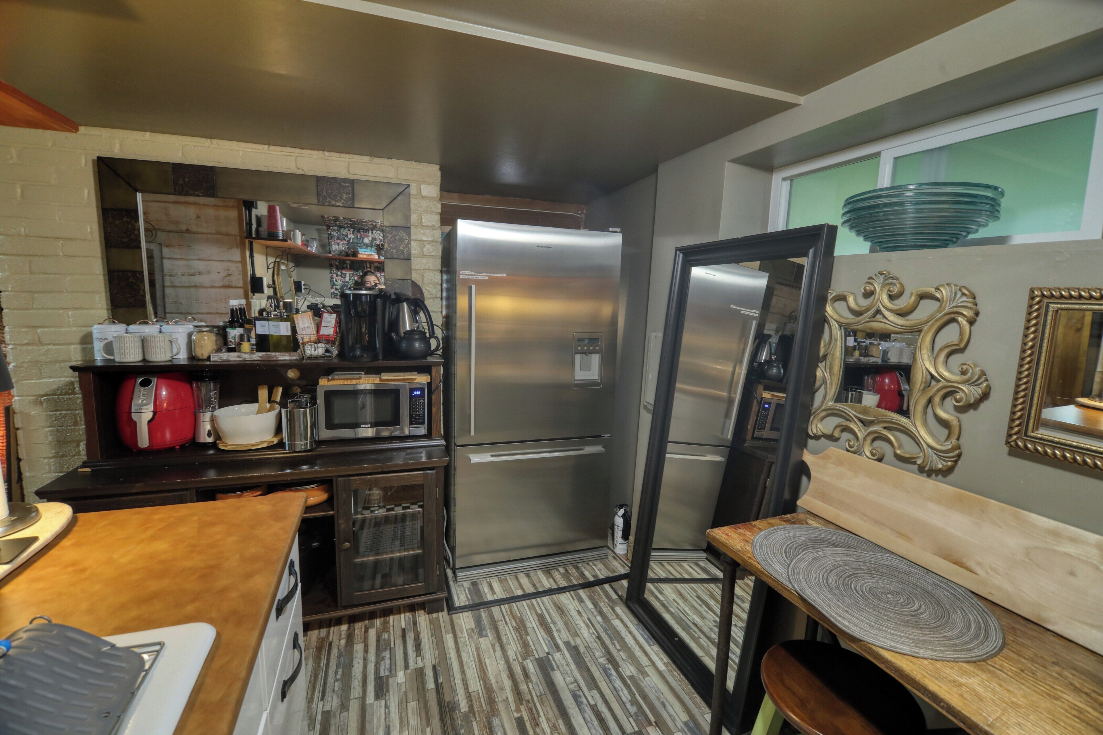 A compact kitchen with a refrigerator, microwave, and coffee maker. There is a wooden table with a bench and a decorative mirror on the wall.