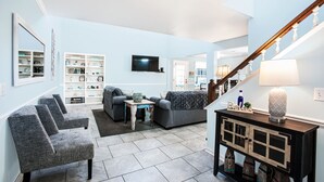 A spacious living room with a grey sofa, two armchairs, a coffee table, and a TV mounted on the wall. There is a staircase with a wooden railing leading to another level.