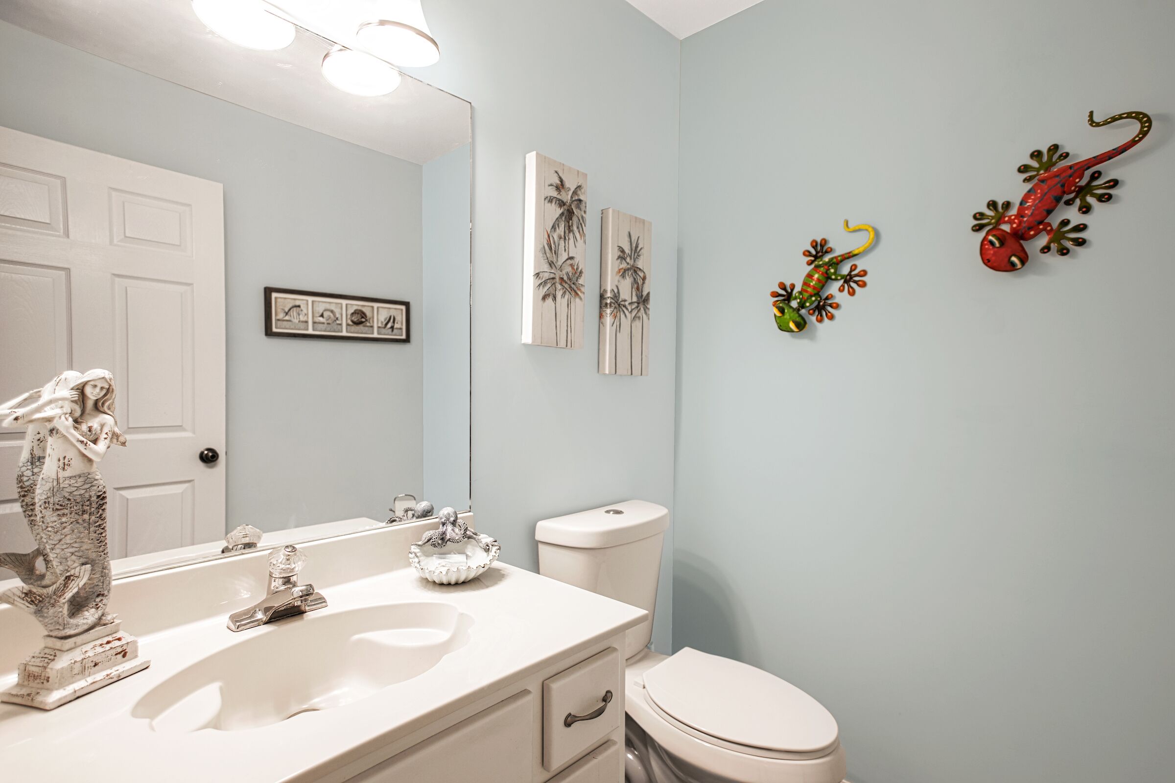 Een badkamer met een wit wastafelmeubel, toilet en spiegel. Twee kunstwerken aan de muur met afbeeldingen van palmbomen.