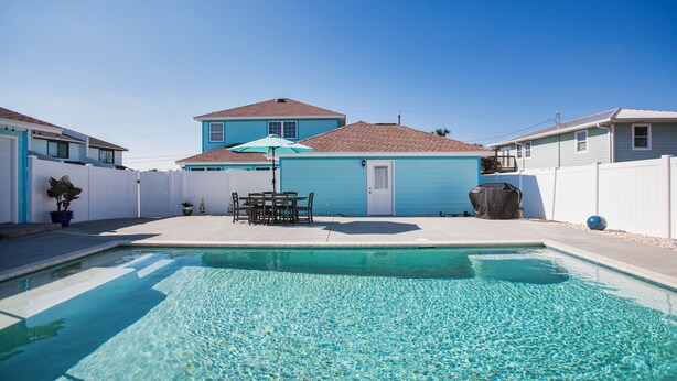 A swimming pool with a patio area, a table with chairs, and a blue house in the background.