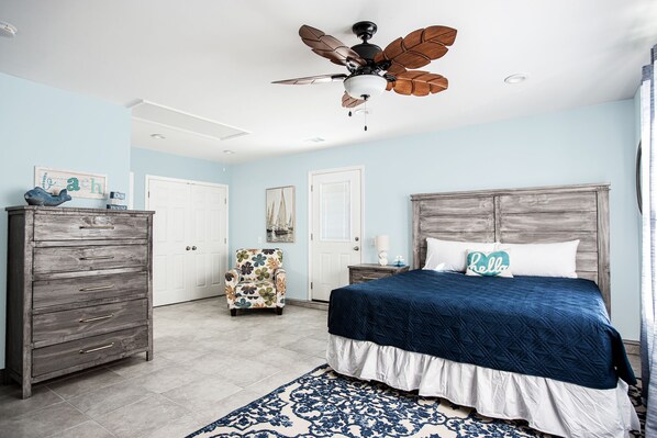 Een slaapkamer met een houten dressoir, een bed met een blauwe sprei, een plafondventilator en een fauteuil met bloemenprint.