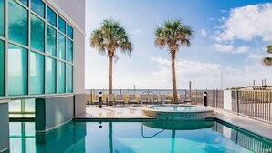 Ein heller, überdachter Pool mit klarem, blauem Wasser, umgeben von Liegestühlen und Palmen, mit Blick auf den Himmel und ein Gebäude mit großen Fenstern.