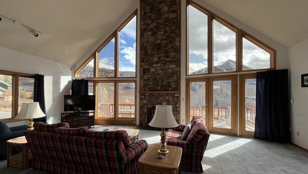 Un salón comedor amplio con techo alto, ventanas grandes que ofrecen vistas a las montañas, chimenea de piedra y mobiliario a cuadros.