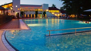 A swimming pool with a tiled deck, a building with large windows in the background, and a railing around the pool. - Aparthotel am Sonnenhuegel (Bad Birnbach)