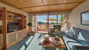 A cozy living room with a wooden ceiling, a television on a wooden cabinet, a sofa, a table with a bowl of fruit, and a view of the outdoors through large windows. - Aparthotel am Sonnenhuegel (Bad Birnbach)