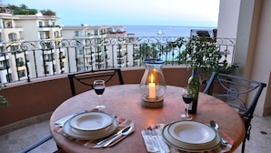 A round table set for two with plates, cutlery, and wine glasses, a lit candle, and a bottle of wine on a balcony overlooking the sea.