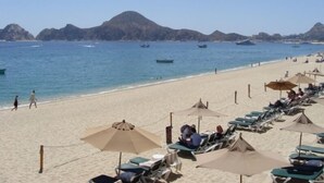 A beach with rows of sun loungers and umbrellas, a clear blue sea, and distant rocky islands.