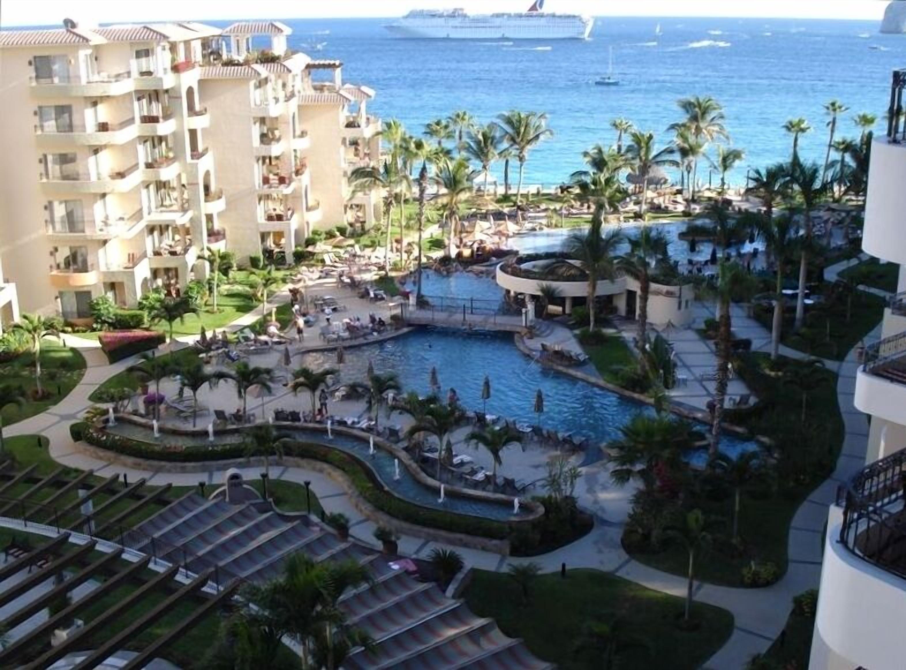 A resort with multiple swimming pools, palm trees, and a view of the ocean and cruise ship.