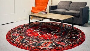Un salon avec un canapé gris, un fauteuil orange, une table basse en bois, un tapis rouge à motifs, un ventilateur de plafond et deux lampes fixées au mur.