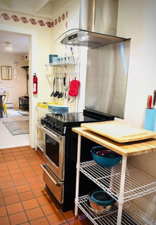 A kitchen with a stove, oven, and a stainless steel range hood. There is a shelf with kitchen utensils and a cutting board on a stand. - Catskill Cabin Duplex Apartment (Shandaken)
