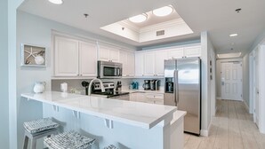 A modern kitchen with white cabinetry, a central island, and stainless steel appliances. - Seawind 1007 | Updated condo with perfect beach view and walk to dine, shop, more! (Gulf Shores)