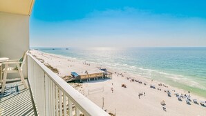 A beach view from a balcony with a clear sky, a pier in the distance, and a sandy beach with umbrellas and people. - Seawind 1007 | Updated condo with perfect beach view and walk to dine, shop, more! (Gulf Shores)