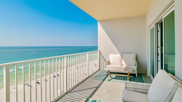 A balcony with a view of the beach and ocean, a white sofa, and a wicker chair. - Seawind 1007 | Updated condo with perfect beach view and walk to dine, shop, more! (Gulf Shores)