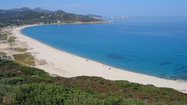 Una playa costera con arena, aguas azules cristalinas y un paisaje montañoso al fondo.