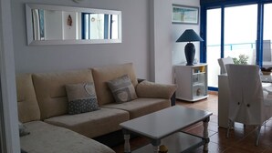 Ett vardagsrum med en beige soffa, ett soffbord och en lampa. Det finns en balkong med en stol och ett bord.