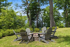 Een vuurkorf met vier Adirondack-stoelen eromheen, omgeven door weelderige begroeiing en bomen.