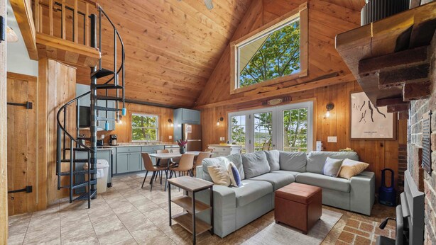 A spacious living area with a large sofa, a coffee table, and a fireplace. A spiral staircase leads to an upper level. - Stowaway - A Private Lakefront Experience With Over 100ft Of Shoreline! (Lodi)