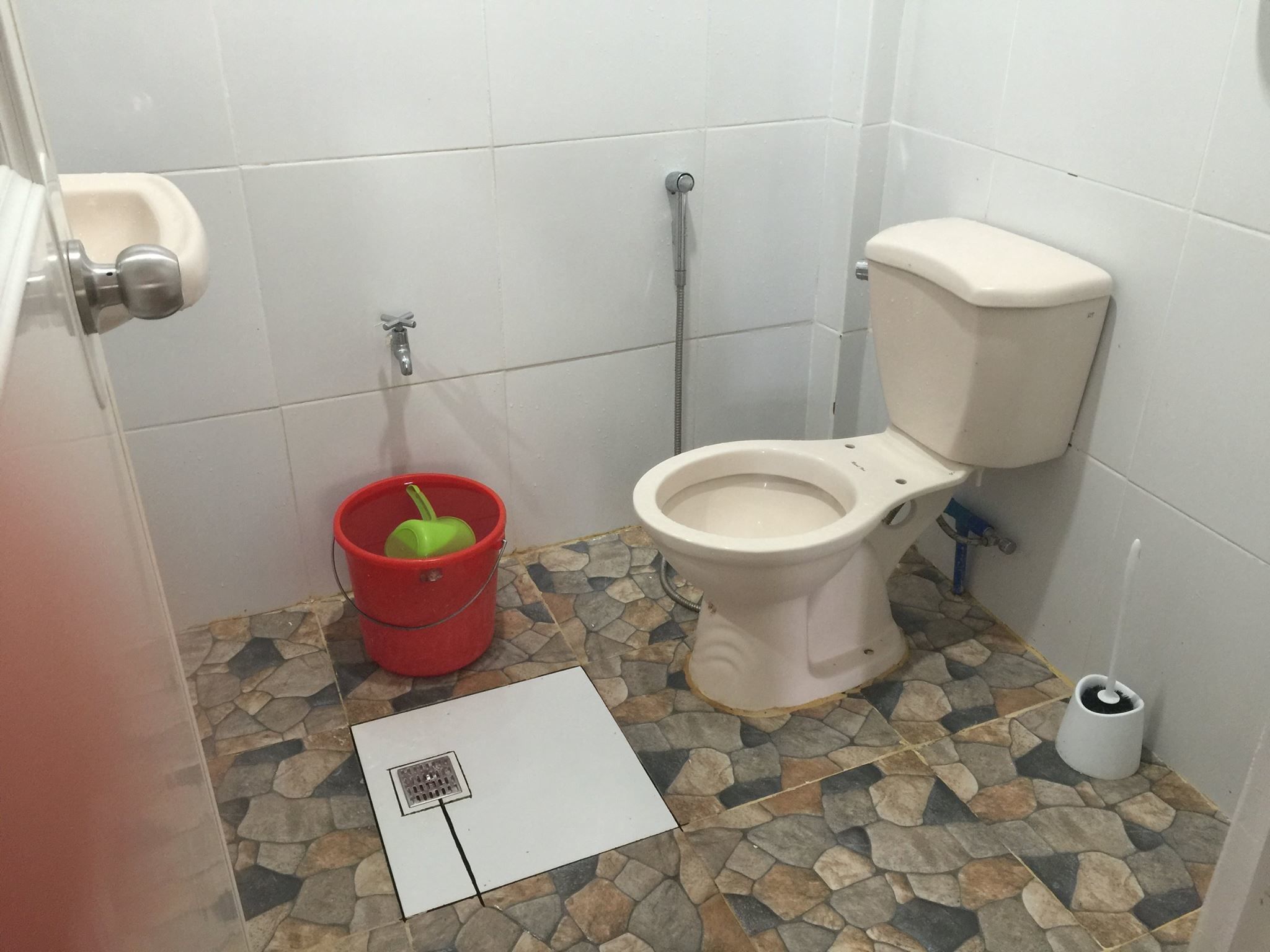 A bathroom with a toilet, a red bucket, and a white floor drain.