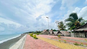 A beachside walkway with a paved path, palm trees, and thatched-roof huts.