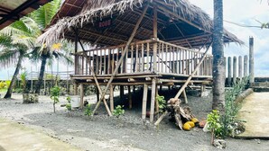 A rustic wooden structure with a thatched roof, labeled "ROSE," surrounded by palm trees and a dirt ground.