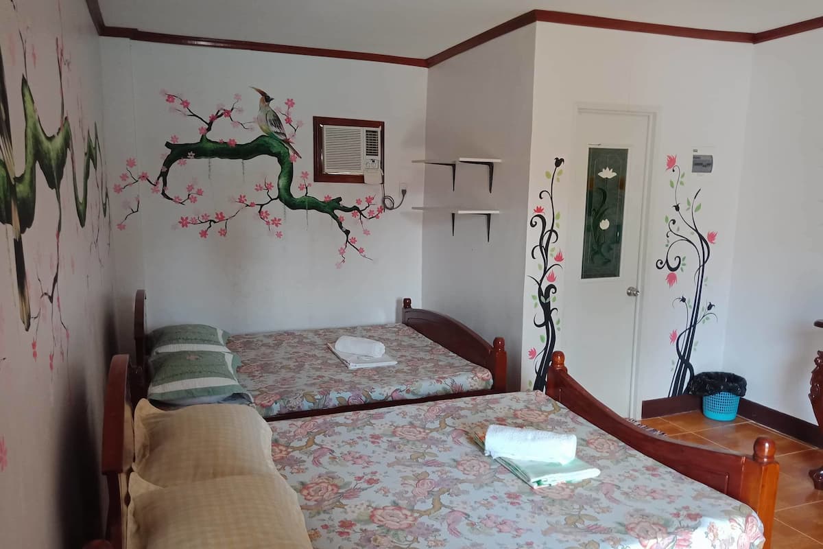 A room with two beds, floral wall art, a door with a decorative panel, and a small trash bin.